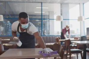 Two men wearing black aprons and gloves while cleaning and disinfecting tables at a commercial restaurant