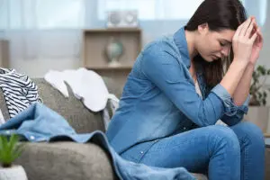A young woman sitting on a messy couch with clothes lying around and holding her head in her hands because she does not want to clean in Springfield, IL.