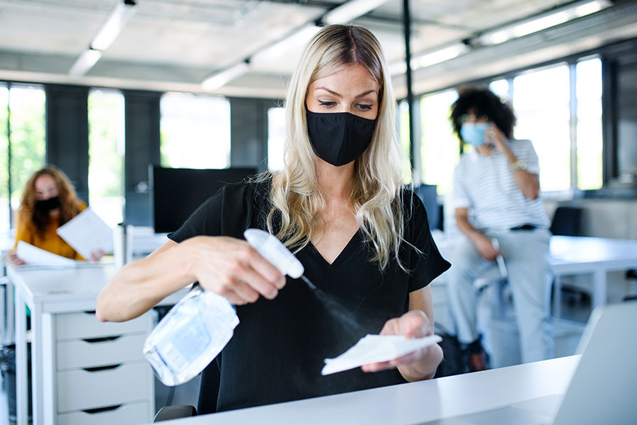Woman in a black t-shirt with a black mask spraying a cleaning on a white rag to clean work desk
