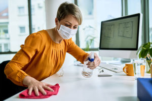A woman wearing an orange shirt with a white face mask spraying down a white office desk with a computer at her job