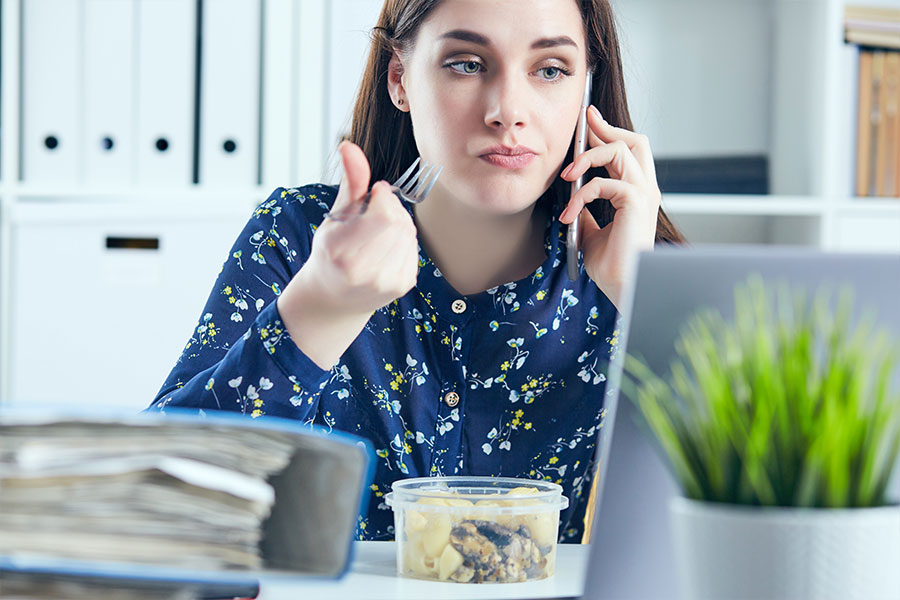 Woman in a blue floral long sleeve shirt eating a leftover meal at her office desk and making a mess near work files and computer