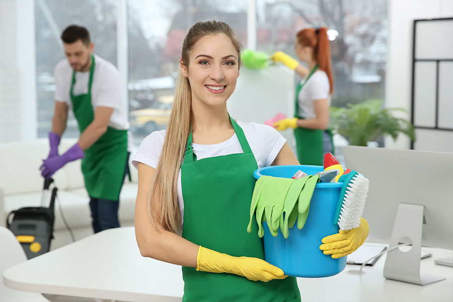 Woman with long blonde hair in a white t-shirt and green apron cleaning an office space with two coworkers and a blue bucket with cleaning supplies