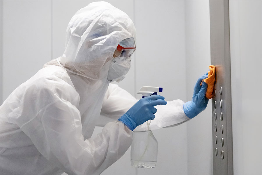 Person in a white hazard suit and blue gloves disinfecting a panel of elevator buttons with chemicals and an orange rag