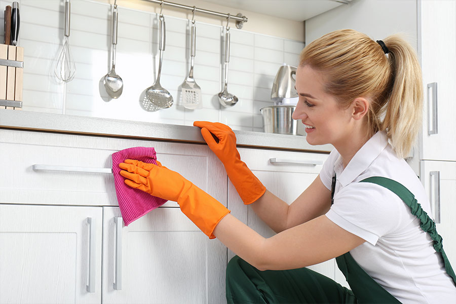 Cleaning woman in a white t-shirt, green overalls, and orange gloves holding a pink washrag and wiping down the cabinets of a residential kitchen for the holidays in Springfield, IL.