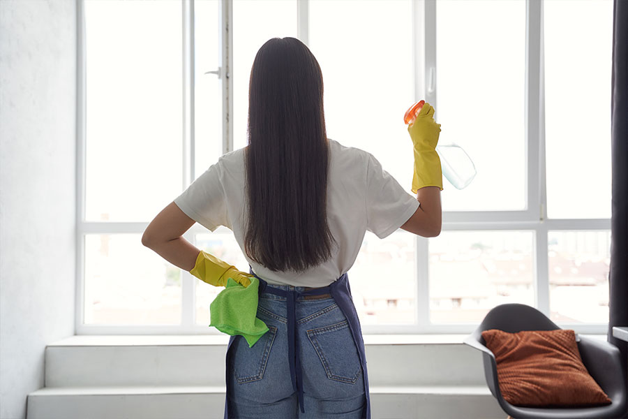 Professional cleaning woman in a white t-shirt, jeans, a blue apron, and yellow gloves spraying disinfectant in a residential home.