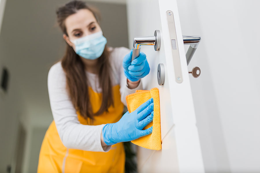 A professional maid in a long white sleeve shirt, yellow apron, and blue gloves wiping down a door handle in a small business in Springfield, IL.