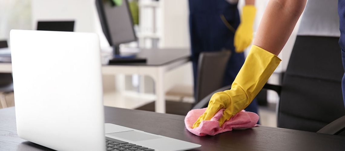 Two cleaning professionals in blue overalls and yellow gloves cleaning office desks and computers with pink and green washcloths