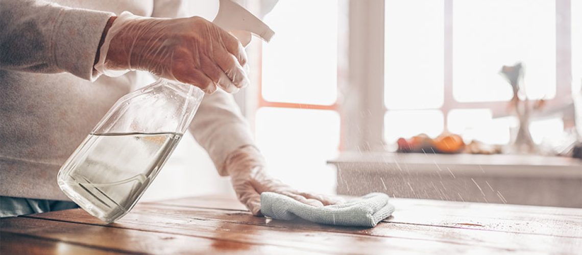 Person in a white shirt with gloves wiping down a wooden table with disinfectant to prevent COVID-19