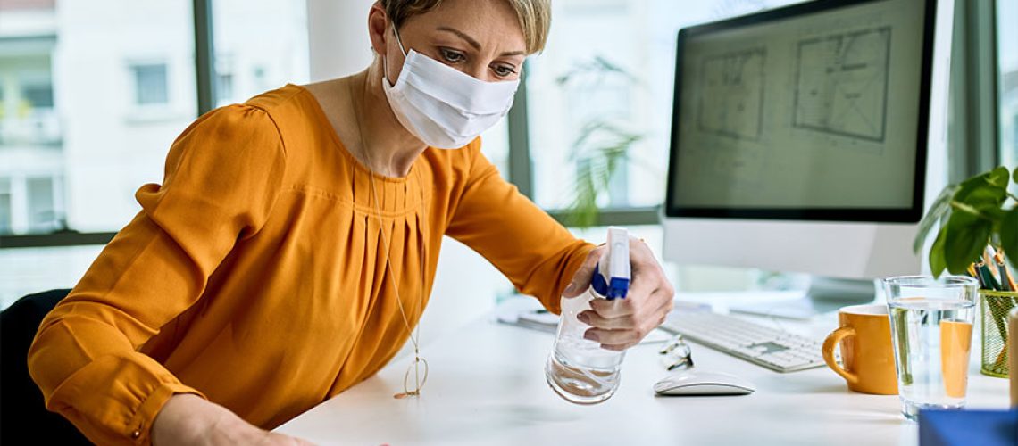 A woman wearing an orange shirt with a white face mask spraying down a white office desk with a computer at her job